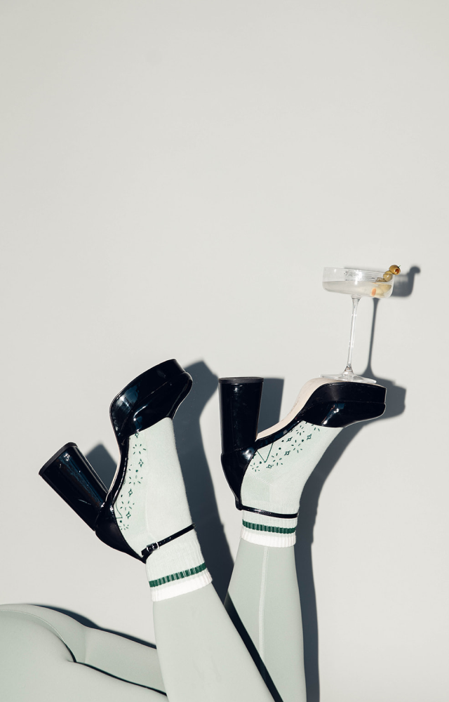 A dirty martini balancing on a high heel with sage green Pilates socks underneath to show the functional grip and design on the bottoms.