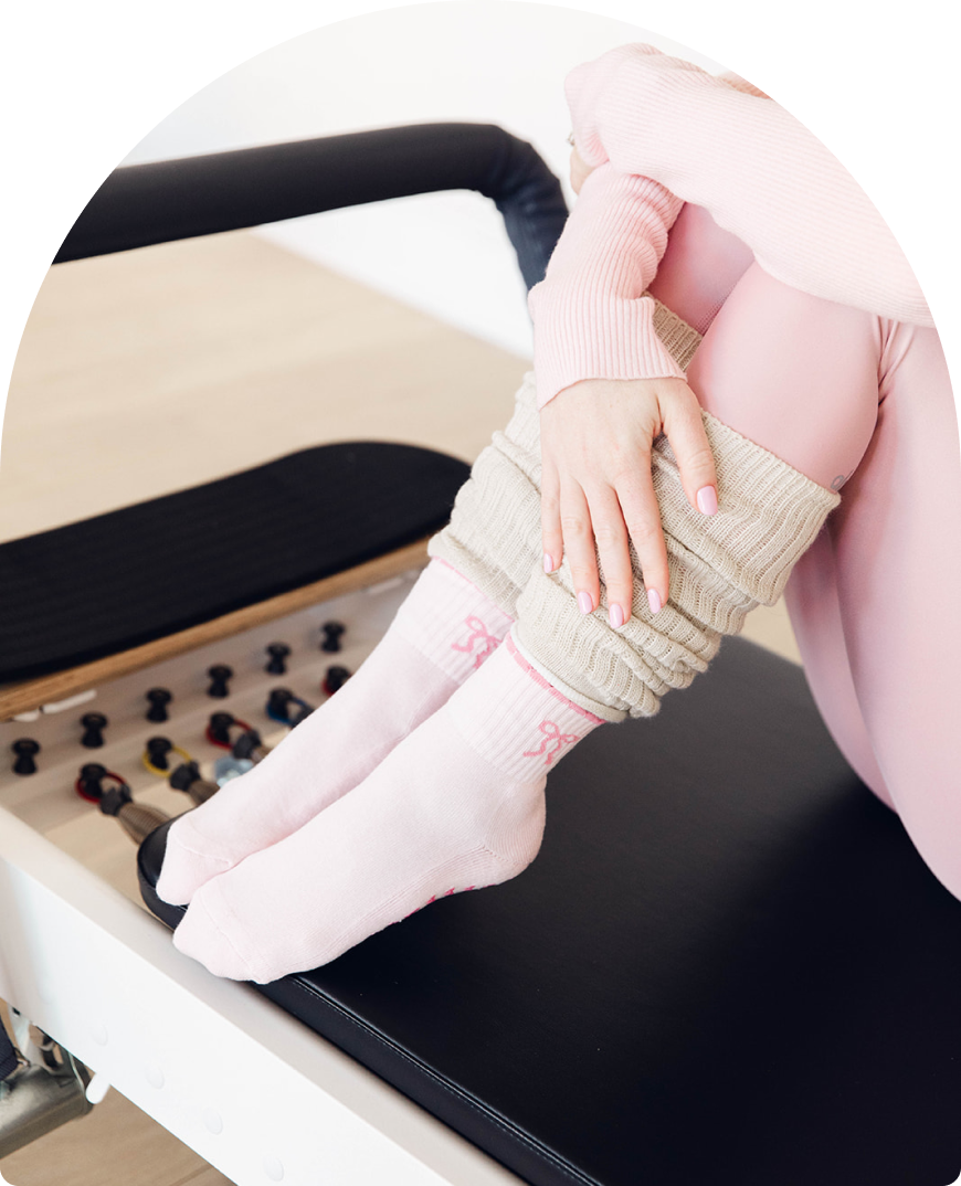 Girl in pink leggings, pink bow Pilates socks and leg warmers sitting on a Pilates reformer before class. 