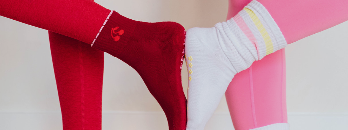 A foot wearing red cherry Pilates socks touching another foot wearing a white retro Pilates sock showcasing their functional grips and designs. 
