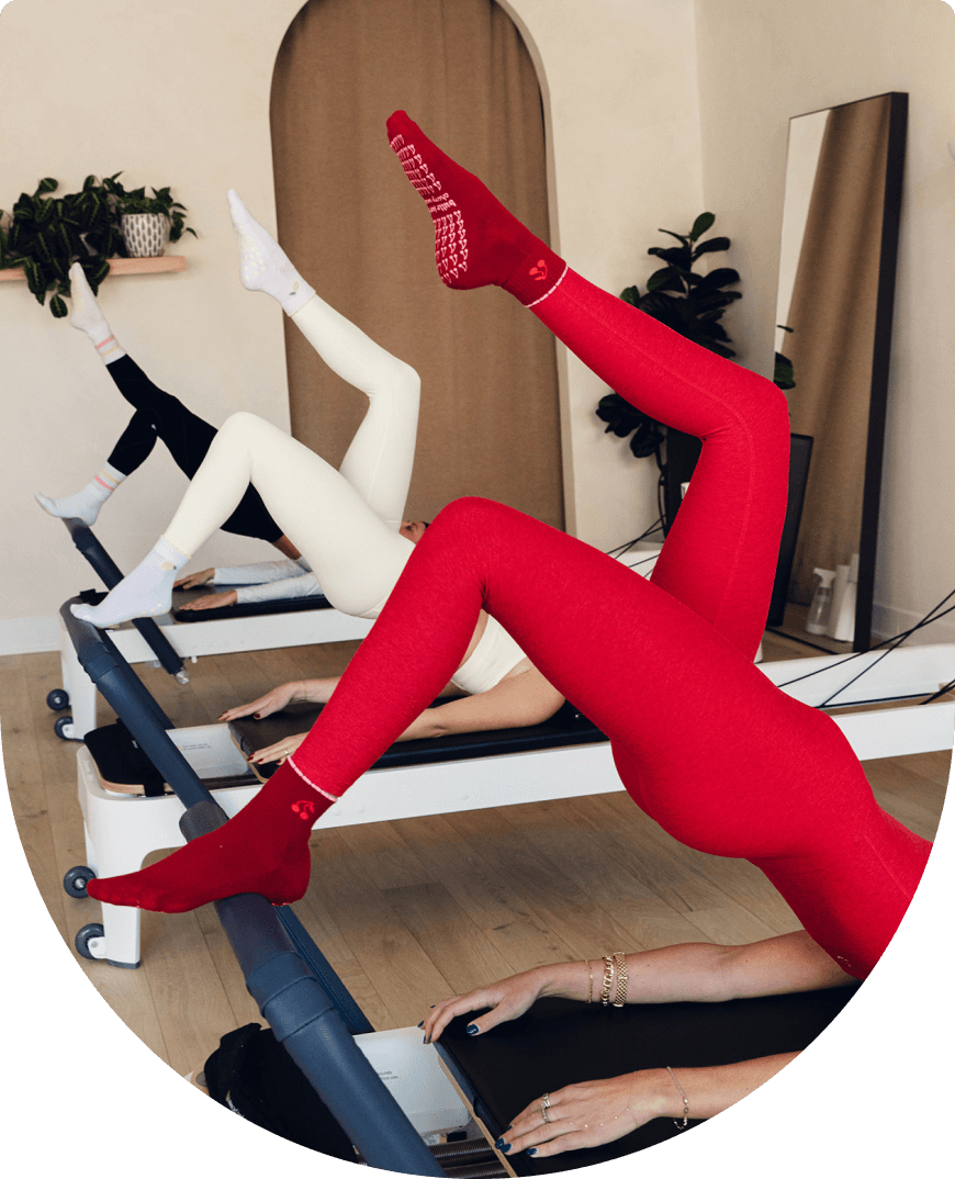 3 girls in different colored outfits to match their unique Pilates socks, doing a bridge pose on a Pilates reformer during class. 
